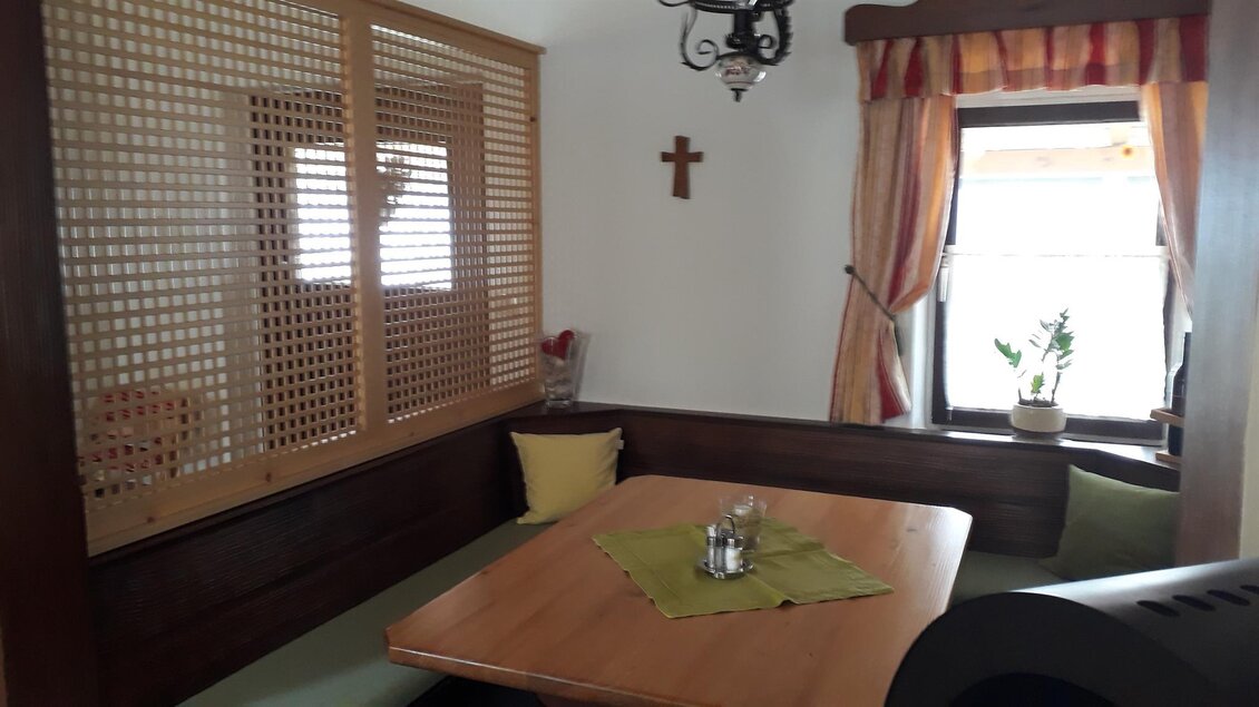 Ein gemütlicher Raum mit einem Esstisch und einer Bank. An der Wand hängt ein Kreuz, und ein Fenster lässt viel Licht herein. | © Andreas Wutti | Weingut Buschenschank Wutti