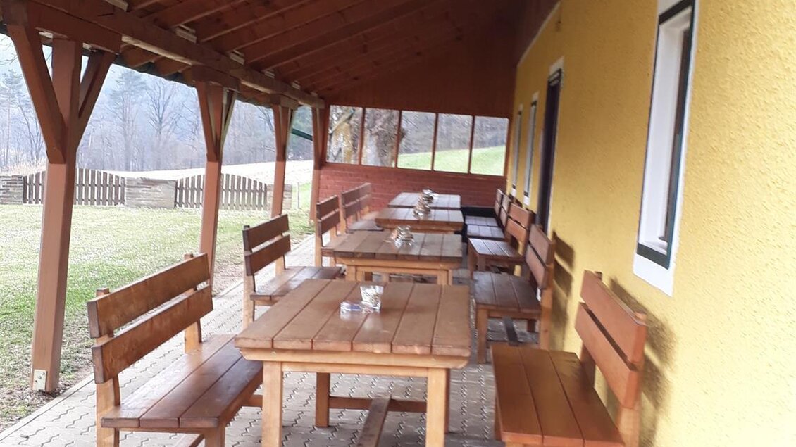 Ein überdachter Außenbereich mit Holztischen und Bänken. Die Wände sind gelb gestrichen und es gibt einen Blick auf die grüne Umgebung. | © Andreas Wutti | Weingut Buschenschank Wutti