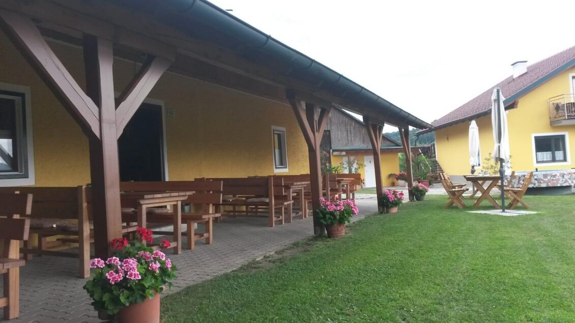 Ein gemütlicher Außenbereich mit Holzmöbeln und bunten Blumen. Im Hintergrund befindet sich ein gelbes Haus und eine grüne Wiese. | © Andreas Wutti | Weingut Buschenschank Wutti