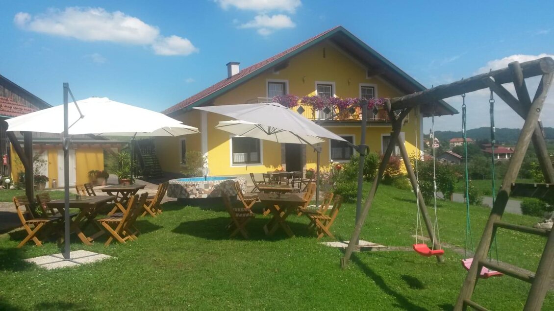 Ein gelbes Haus umgeben von einem grünen Garten. Es gibt Tische mit Sonnenschirmen und eine Schaukel im Außenbereich. | © Andreas Wutti | Weingut Buschenschank Wutti