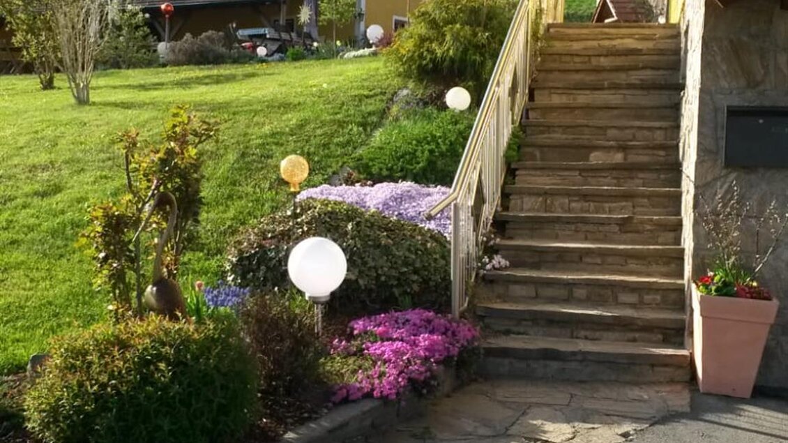 Eine schöne Gartenlandschaft mit Treppen und bunten Blumen. Im Hintergrund sind Gebäude und ein klarer Himmel zu sehen. | © Andreas Wutti | Weingut Buschenschank Wutti
