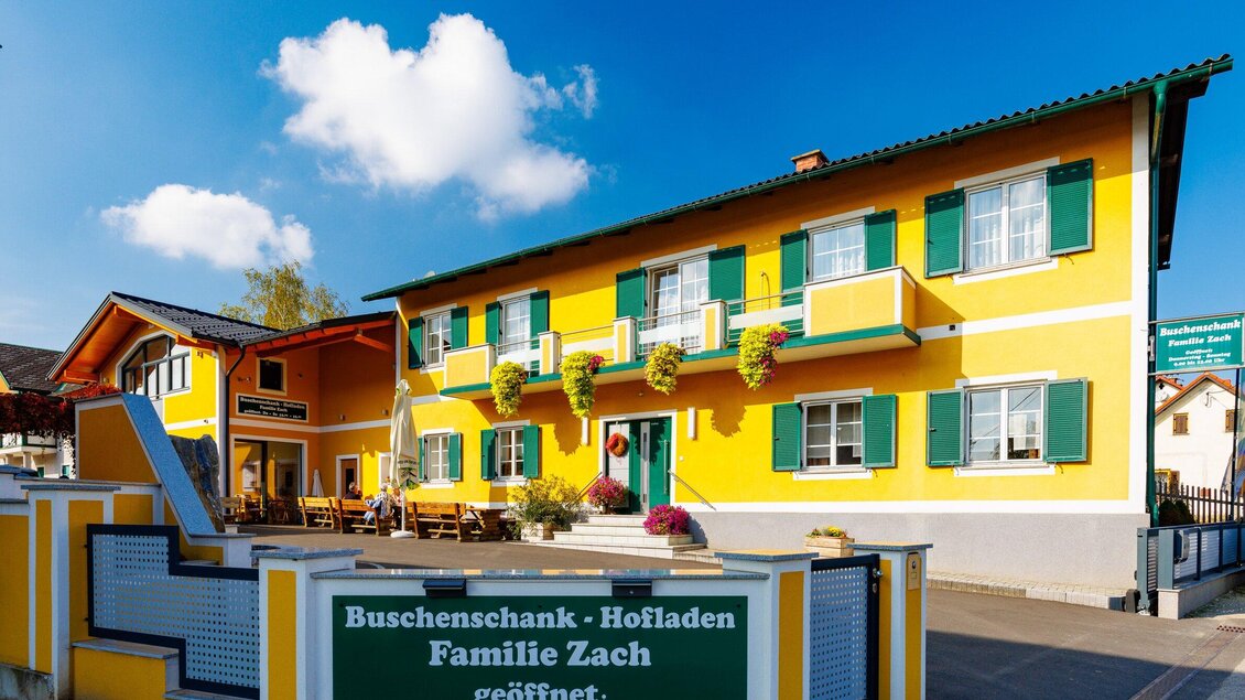 Ein hellgelbes Gebäude mit grünen Fensterläden und einem einladenden Außensitzbereich. Das Schild weist auf den Buscheneschank und Hofladen der Familie Zach hin. | © TV Region Graz - Harry Schiffer