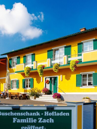 Ein hellgelbes Gebäude mit grünen Fensterläden und einem einladenden Außensitzbereich. Das Schild weist auf den Buscheneschank und Hofladen der Familie Zach hin. | © TV Region Graz - Harry Schiffer