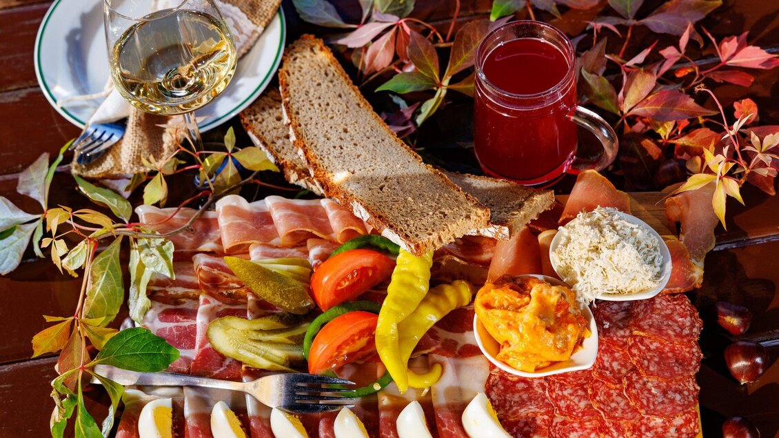 Ein reichhaltiges Frühstücksbuffet mit verschiedenen Wurstsorten, frischem Gemüse, Brot und Aufstrichen. Daneben stehen ein Glas Wein und ein Getränk auf einem rustikalen Tisch mit herbstlichen Blättern. | © TV Region Graz - Harry Schiffer