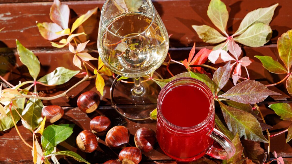 Zwei Getränke, ein Glas Weißwein und ein Becher mit rotem Getränk, liegen auf einem Tisch. Umgeben sind sie von buntem Herbstlaub und Kastanien. | © TV Region Graz - Harry Schiffer