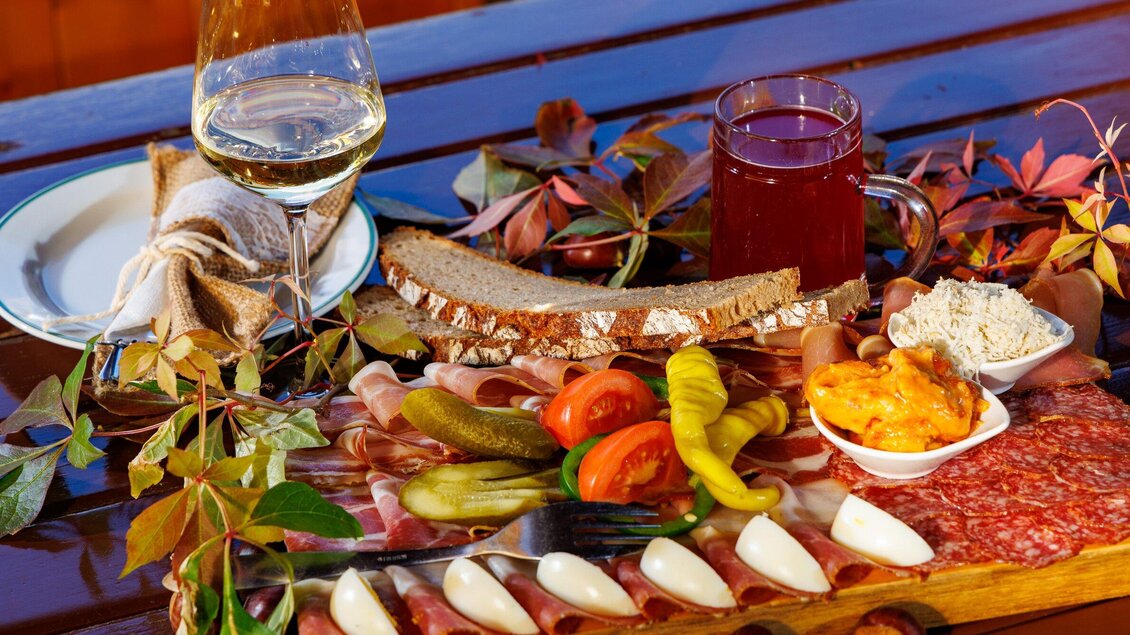 Ein schön angerichteter Teller mit Wurst, Käse, eingelegtem Gemüse und Brot. Dazu ein Glas Weißwein und ein Getränk in einer Tasse, umgeben von bunten Blättern. | © TV Region Graz - Harry Schiffer