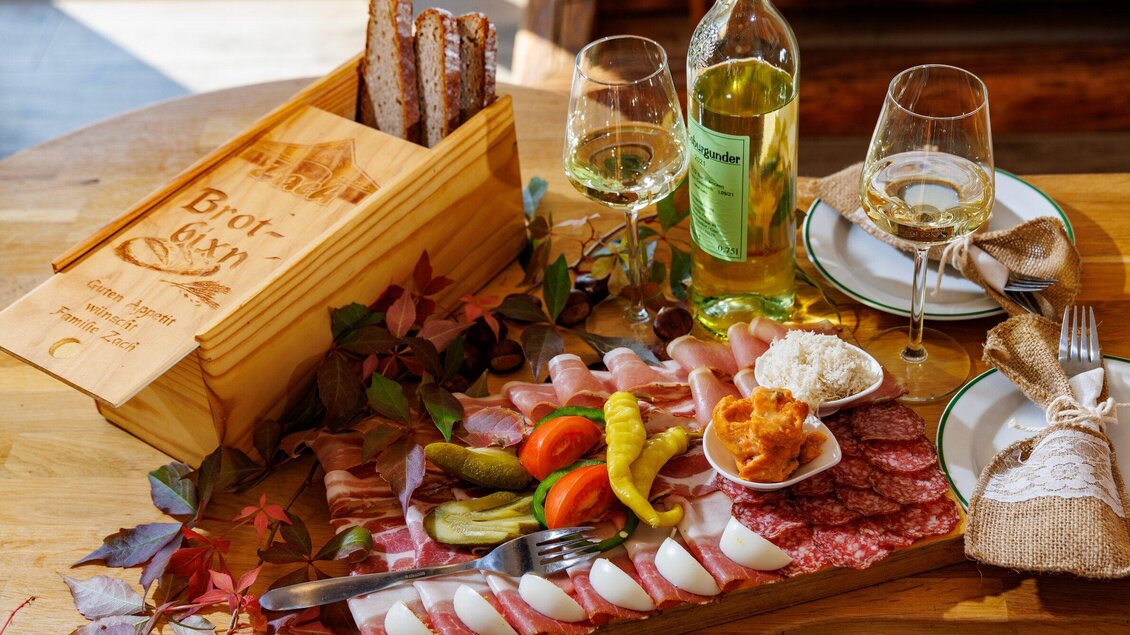Ein reichhaltiges Brett mit verschiedenen Wurst- und Käsesorten, eingelegtem Gemüse und Brot. Dazu zwei Gläser Weißwein und dekorative Blätter auf dem Tisch. | © TV Region Graz - Harry Schiffer