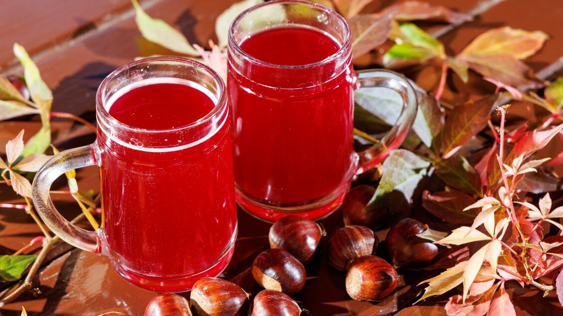 Zwei Gläser mit rotem Getränk stehen auf einem Tisch, umgeben von buntem Herbstlaub und Kastanien. Die warme Farbgebung vermittelt eine gemütliche Herbststimmung. | © TV Region Graz - Harry Schiffer