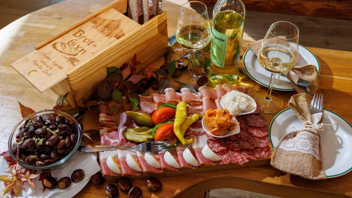 Ein reichhaltiges Käse- und Wurstbrett mit verschiedenen Beilagen, umgeben von Wein und Glaswaren. Deftige Snacks und eine rustikale Präsentation schaffen eine einladende Atmosphäre. | © TV Region Graz - Harry Schiffer