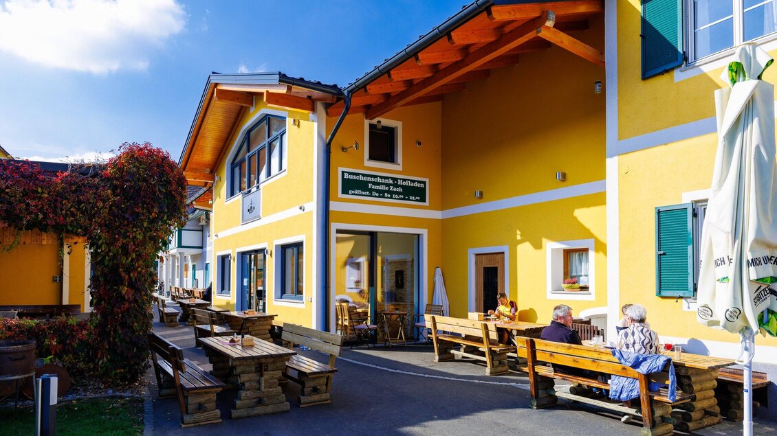 Ein gelbes Gebäude mit einer einladenden Außenterrasse. Gäste sitzen auf Holzbank und genießen die sonnige Atmosphäre. | © TV Region Graz - Harry Schiffer