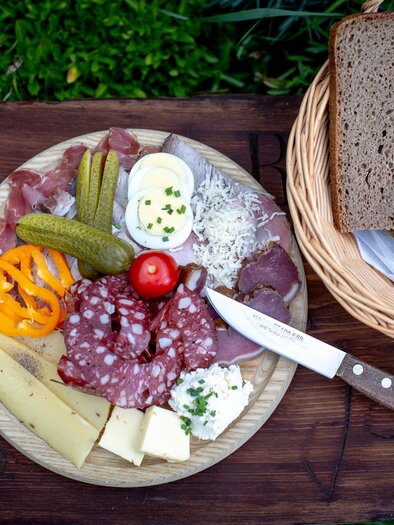 A plate with various types of sausages and cheeses, pickled cucumbers, and a small salad. Next to it is a basket with fresh bread. | © TV Murau
