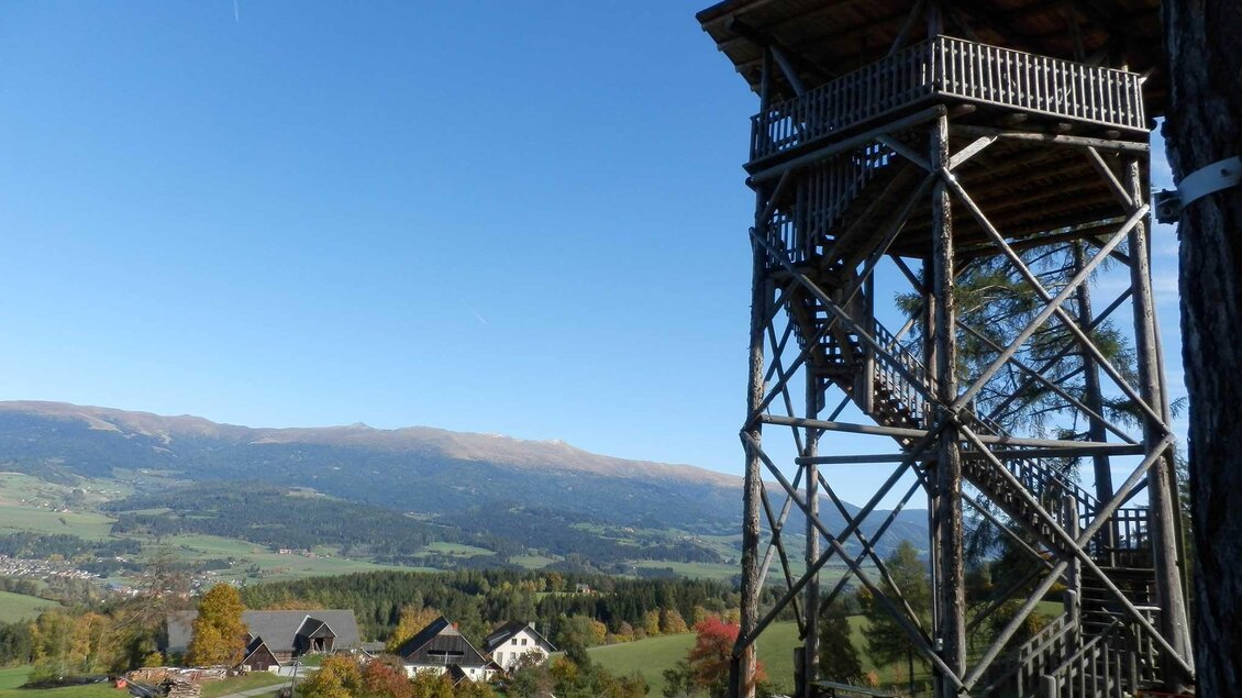 Ein hölzernes Aussichts-Turm steht auf einer Wiese mit Blick auf das Tal. Im Hintergrund sind sanfte Hügel und einige Häuser zu sehen. | © Buschenschank Zeischgl