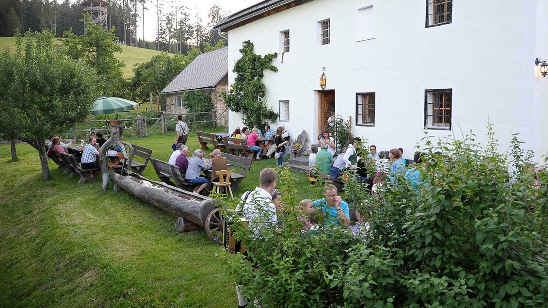 Eine gemütliche Außenbereich mit vielen Gästen, die an Tischen sitzen und essen. Im Hintergrund ist ein rustikales Haus und grüne Bäume zu sehen. | © Buschenschank Zeischgl