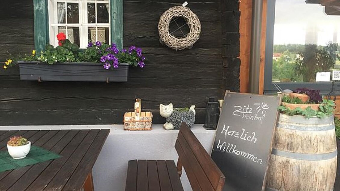 Ein einladender Außenbereich eines Hauses mit Holzwänden. Auf dem Tisch steht eine kleine Pflanze und eine Tafel mit dem Schriftzug "Herzlich Willkommen". | © Walter Christian Zitz | Weinhof Zitz