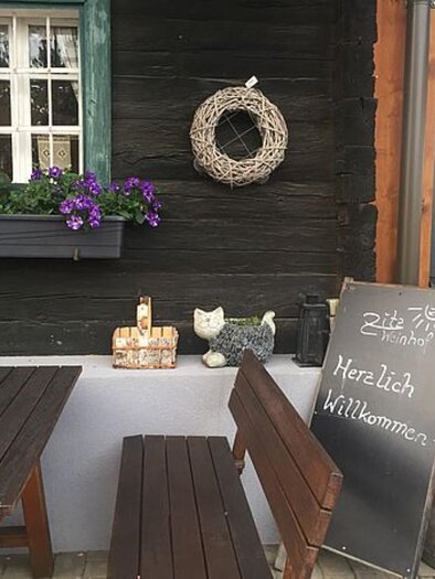 Ein einladender Außenbereich eines Hauses mit Holzwänden. Auf dem Tisch steht eine kleine Pflanze und eine Tafel mit dem Schriftzug "Herzlich Willkommen". | © Walter Christian Zitz | Weinhof Zitz