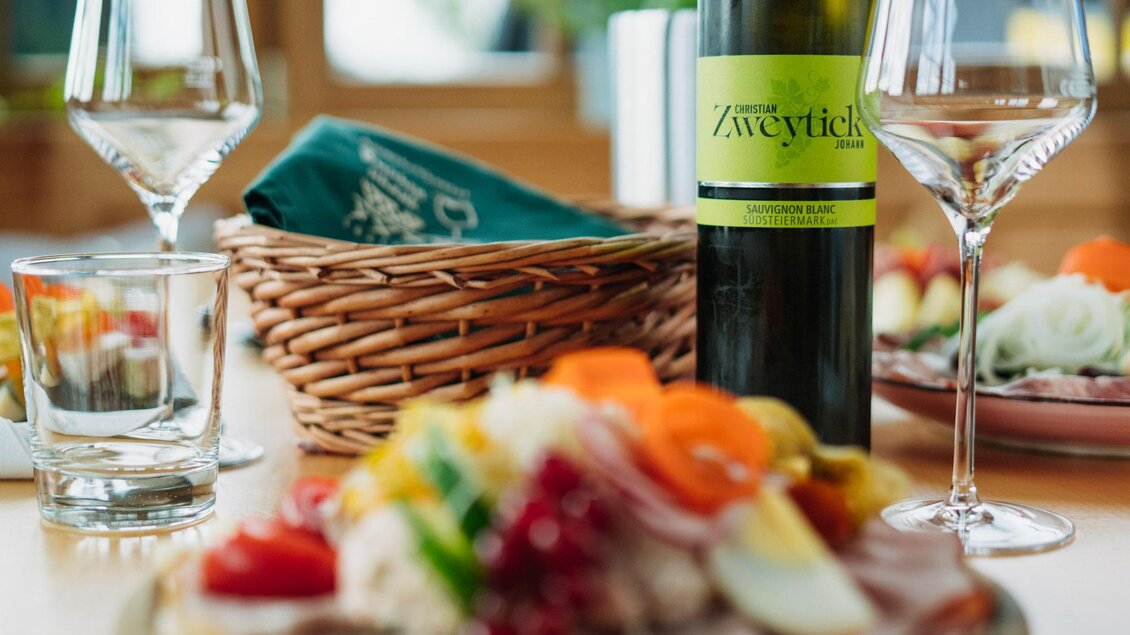 Ein lecker angerichteter Teller mit verschiedenen Speisen und frischen Zutaten. Im Hintergrund stehen ein Wein und Gläser auf dem Tisch. | © Weingut & Buschenschank Zweytick | Fam. Zweytick