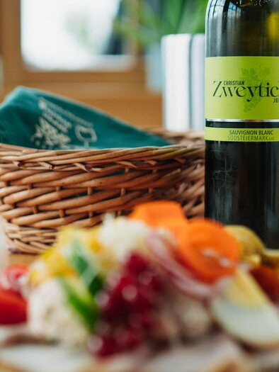 Ein lecker angerichteter Teller mit verschiedenen Speisen und frischen Zutaten. Im Hintergrund stehen ein Wein und Gläser auf dem Tisch. | © Weingut & Buschenschank Zweytick | Fam. Zweytick