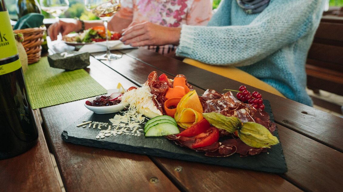 Ein leckerer Teller mit frischem Gemüse, Aufschnitt und Käse auf einem Tisch. Im Hintergrund sind zwei Personen, die gemeinsam speisen. | © Weingut & Buschenschank Zweytick | Fam. Zweytick