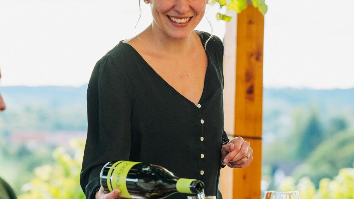 Eine lächelnde Frau gießt Wein in ein Glas an einem schön gedeckten Tisch. Im Hintergrund sind Weinreben und eine malerische Landschaft zu sehen. | © Weingut & Buschenschank Zweytick | Fam. Zweytick