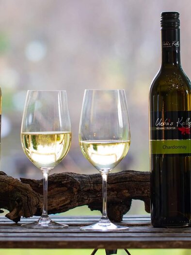 Three bottles of white wine stand next to two glasses filled with wine. In the background, a natural, blurred environment can be seen. | © glaMur