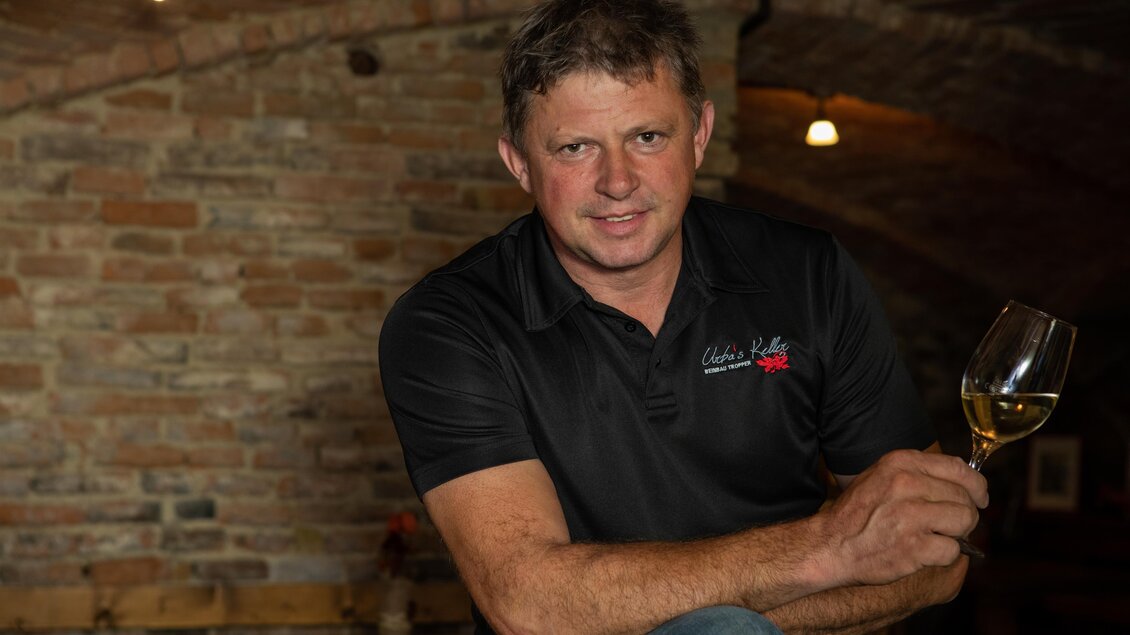 Ein Mann hält ein Weinglas und sitzt in einem rustikalen Keller. Die Steinmauern und das gedämpfte Licht schaffen eine gemütliche Atmosphäre. | © glaMur