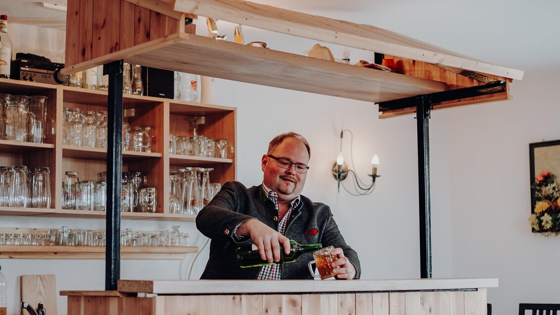 Ein lächelnder Barkeeper schenkt ein Getränk an einer Holzbar aus. Im Hintergrund sind Gläser und eine gemütliche Atmosphäre zu sehen. | © GenussReich