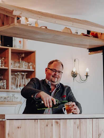 A smiling bartender is serving a drink at a wooden bar. In the background, glasses and a cozy atmosphere can be seen. | © GenussReich