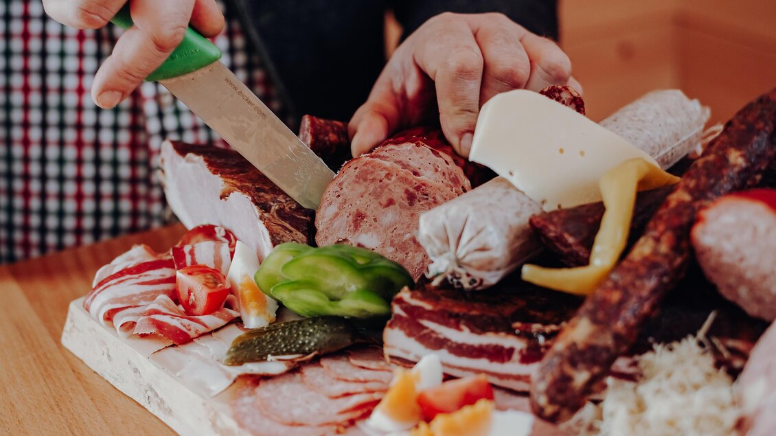 Ein Holzbrett mit verschiedenen Fleischsorten und Käse wird vorbereitet. Jemand schneidet mit einem Messer in die Präsentation. | © GenussReich