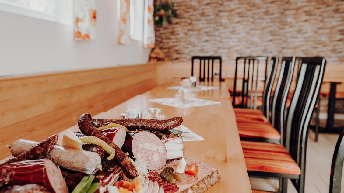 Ein langer Tisch mit Holzoberfläche und mehreren Stühlen steht in einem gemütlichen Raum. Auf dem Tisch liegt eine Auswahl an Wurst und Käse, dekoriert mit Gemüse und Früchten. | © GenussReich