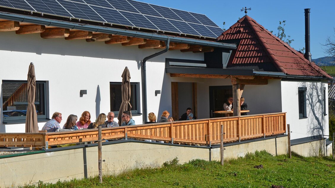 Eine Gruppe von Menschen sitzt an einem Tisch auf einer Terrasse vor einem modernen Haus. Das Gebäude hat Solarpanels auf dem Dach und eine sonnige Umgebung. | © Almenlandblick