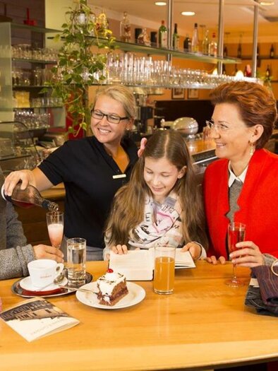 A cozy coffee break in a café with a family and a friendly waitress. They are enjoying drinks and a cake in an inviting atmosphere. | © Cafe im Kaufhaus Hubmann