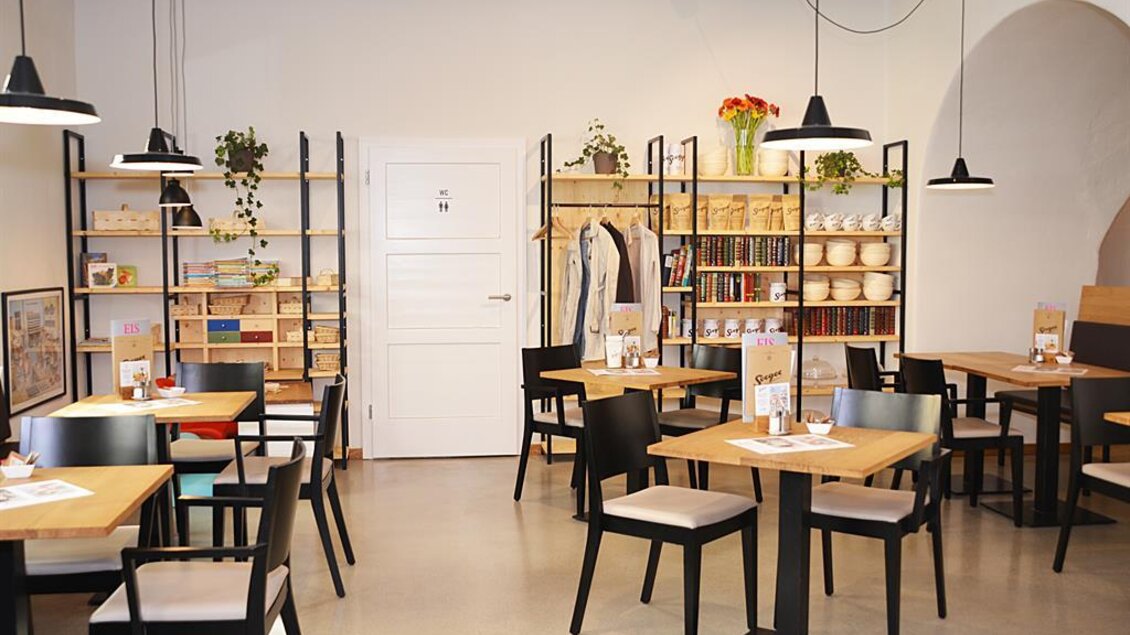 Ein modernes Café mit Holztischen und schwarzen Stühlen. Die Wände sind mit Regalen voller Bücher und Pflanzen dekoriert. | © Werner Krug