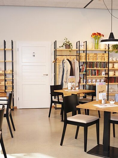 Ein modernes Café mit Holztischen und schwarzen Stühlen. Die Wände sind mit Regalen voller Bücher und Pflanzen dekoriert. | © Werner Krug