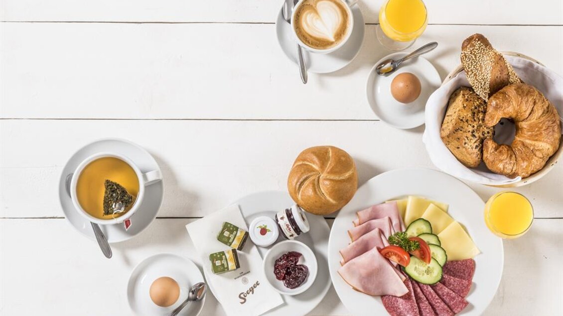 Ein reichhaltiges Frühstück mit Aufschnitt, Käse, Gemüse und frischen Brötchen. Dazu Kaffee, Tee und Orangensaft auf einem weißen Tisch. | © Werner Krug