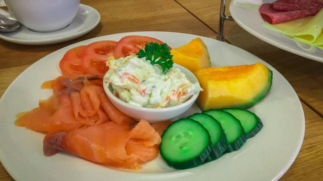 Ein köstlicher Teller mit Räucherlachs, frischen Tomaten, Gurkenscheiben und Melonenstücken. Dazu ein Becher Kaffee im Hintergrund.