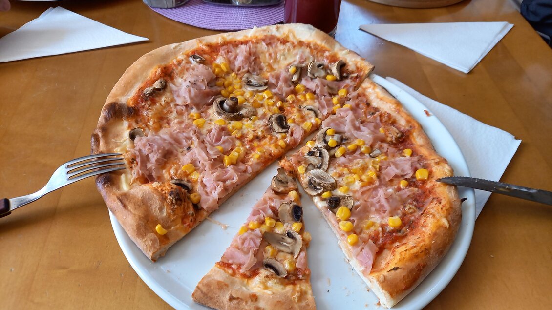 Eine Pizza mit Schinken, Mais und Champignons auf einem weißen Teller. Einige Stücke sind bereits herausgeschnitten und serviert. | ©  Thermen- & Vulkanland