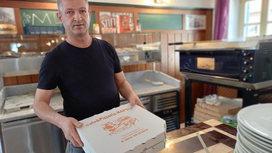 Ein Mann hält eine Pizzaschachtel in einem Restaurant. Im Hintergrund sind eine Pizzateigmaschine und Tische sichtbar. | © Thermen- & Vulkanland
