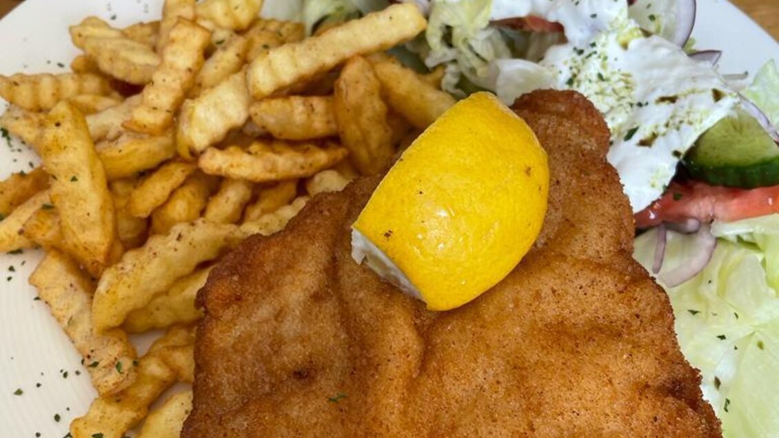 Ein knuspriges Schnitzel mit einer Zitronenscheibe, serviert mit Pommes frites und einem frischen Salat. Das Gericht ist auf einem weißen Teller angerichtet. | © Thermen- & Vulkanland
