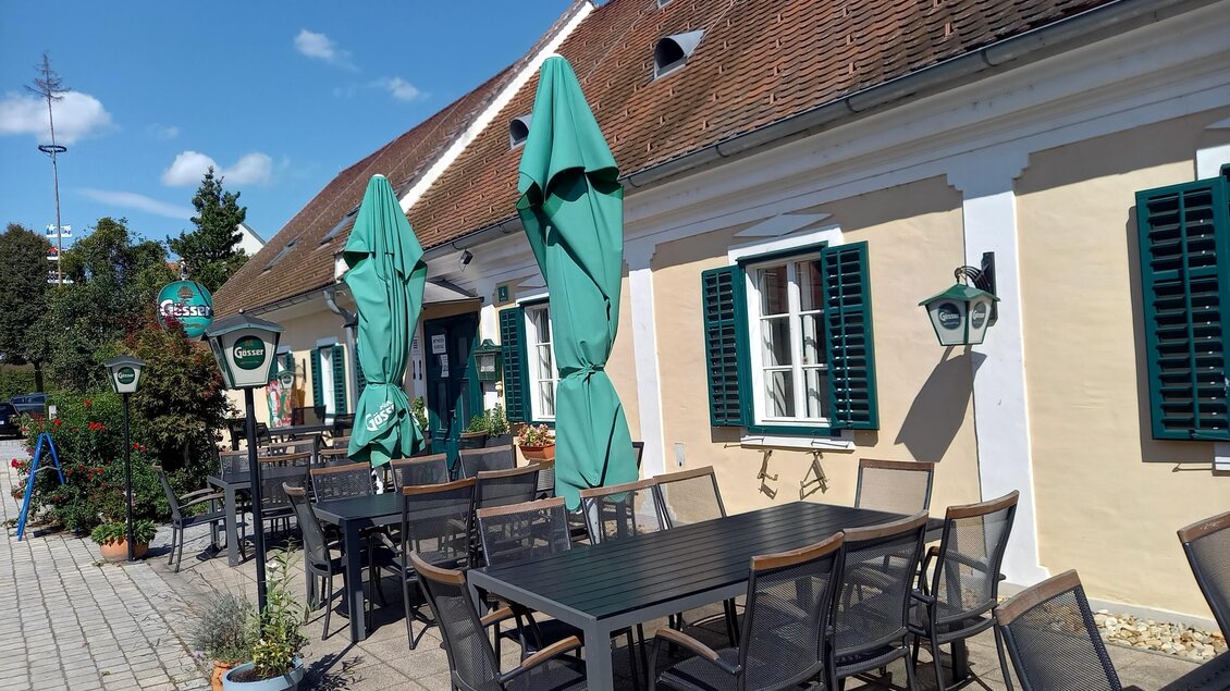 Ein gemütliches Restaurant mit einem Außensitzbereich. Grünes Sonnenschirm und hübsche Fenster verleihen der Umgebung einen einladenden Charakter. | © Thermen- & Vulkanland