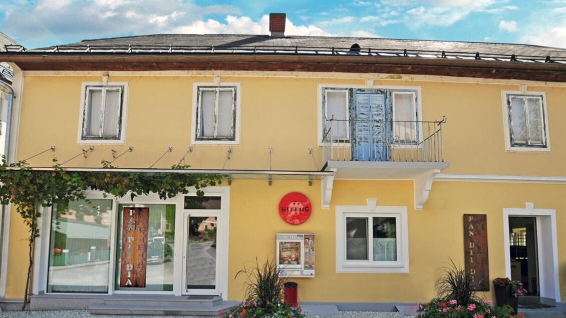 Ein charmantes gelbes Gebäude mit mehreren Fenstern und einer einladenden Eingangstür. Vor dem Haus befinden sich blühende Pflanzen und ein klarer blauer Himmel. | © Sabine Pauritsch