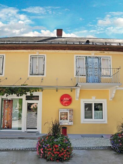 Ein charmantes gelbes Gebäude mit mehreren Fenstern und einer einladenden Eingangstür. Vor dem Haus befinden sich blühende Pflanzen und ein klarer blauer Himmel. | © Sabine Pauritsch
