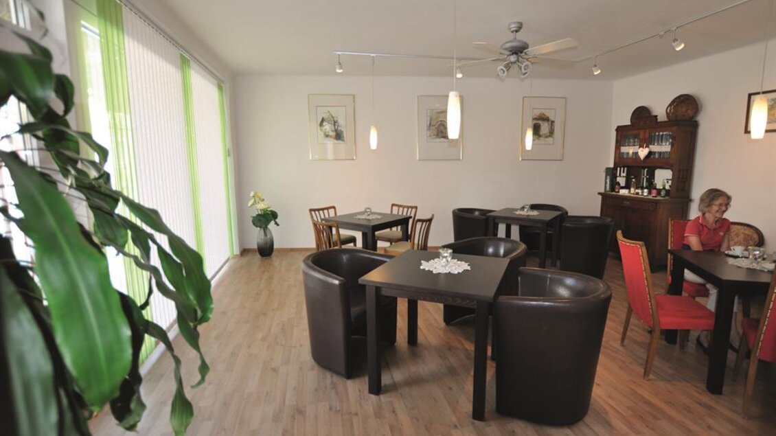 Ein gemütliches Café mit Holztischen und einer warmen Atmosphäre. Im Hintergrund ist eine Frau, die Kaffee trinkt, während die Fenster viel Tageslicht hereinlassen. | © Sabine Pauritsch