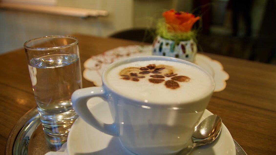 Eine Tasse Kaffee mit einer dekorativen Schaumoberfläche und einigen Kaffeebohnen. Neben der Tasse steht ein Glas Wasser auf einem silbernen Tablett. | © Rainer M. Fürstaller