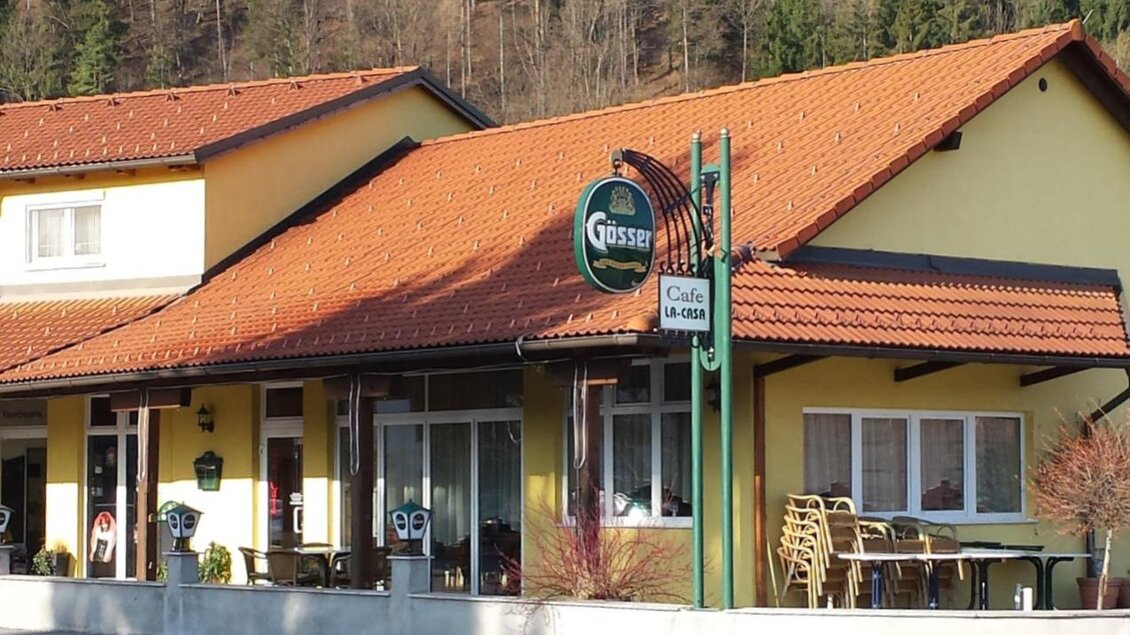 Ein zweigeschossiges, gelbes Gebäude mit einem roten Dach. Vor dem Haus befindet sich ein Café mit Sitzgelegenheiten im Freien. | © Wipfler