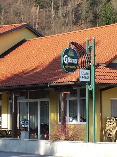 Ein zweigeschossiges, gelbes Gebäude mit einem roten Dach. Vor dem Haus befindet sich ein Café mit Sitzgelegenheiten im Freien. | © Wipfler