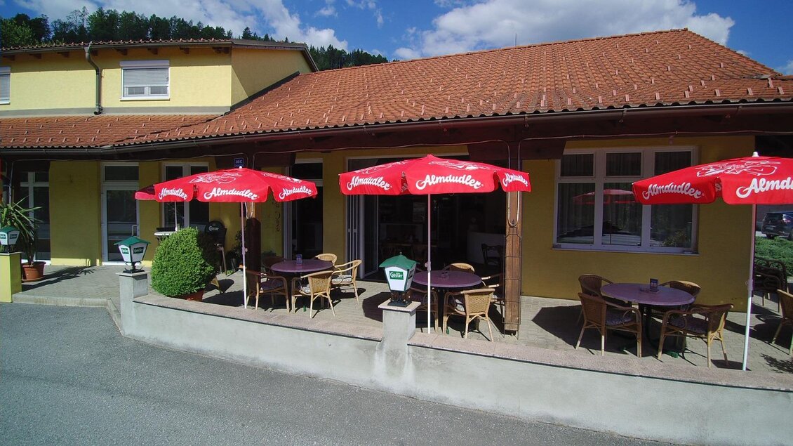 Ein gemütliches Café mit roten Sonnenschirmen und Tischgarnituren im Freien. Die gelbe Fassade und die grüne Umgebung verleihen eine entspannte Atmosphäre. | © La Casa FB