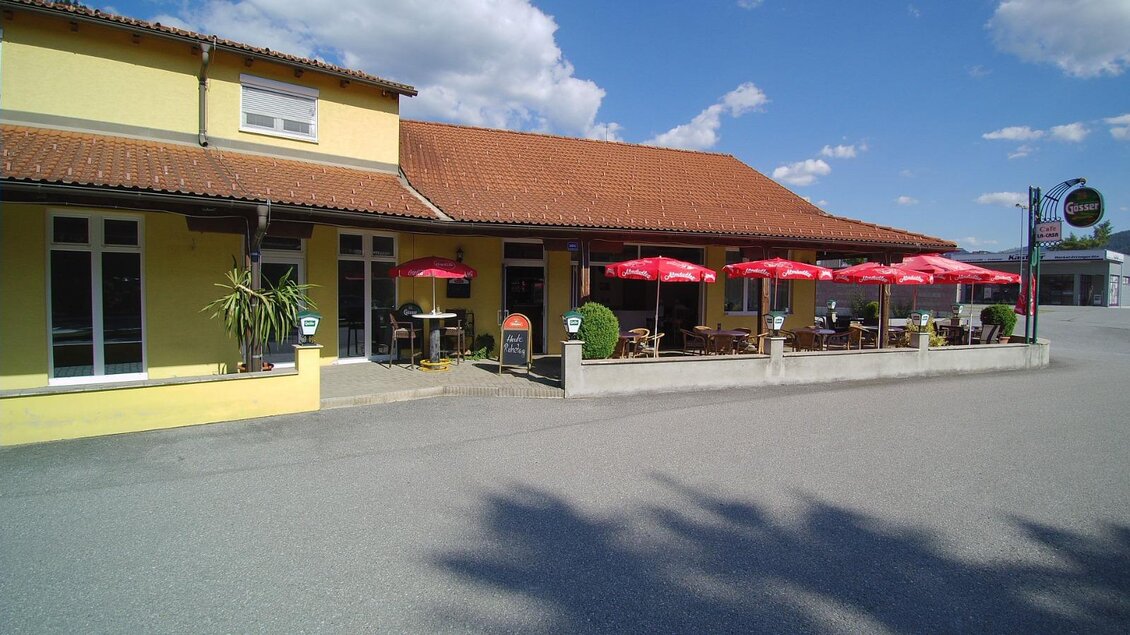 Ein gemütliches Restaurant mit einem hellgelben Außenbereich. Rote Sonnenschirme stehen auf der Terrasse, die am gepflasterten Weg liegt. | © La Casa FB