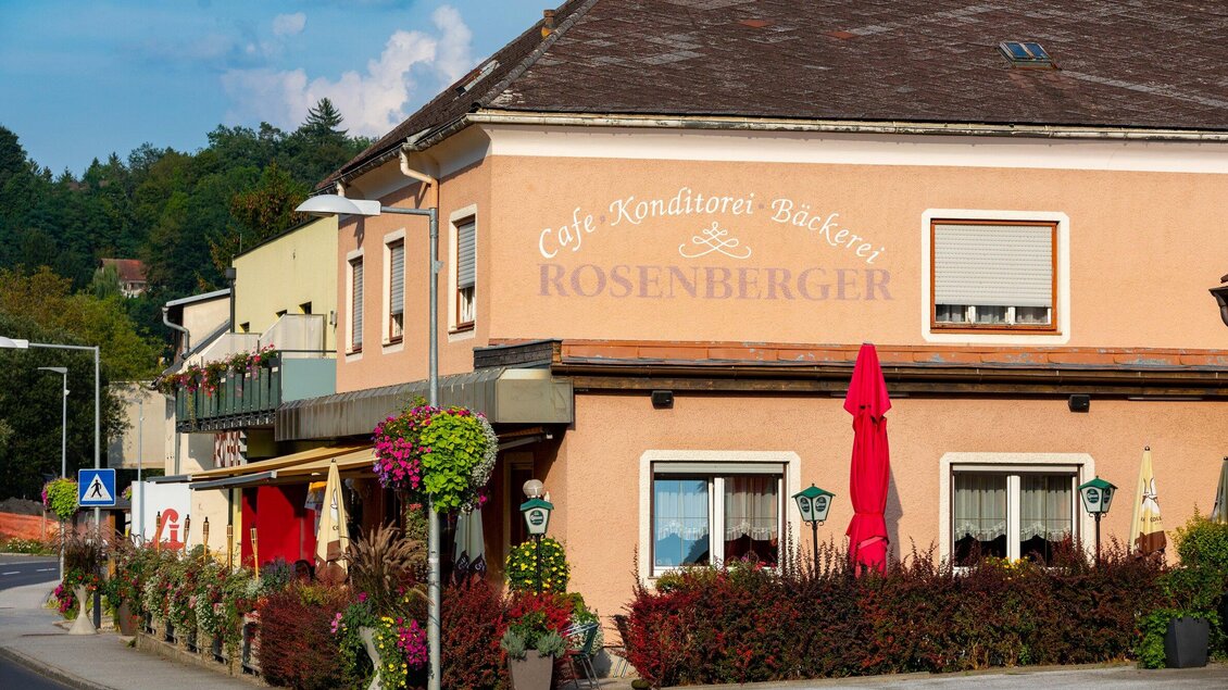 Ein gemütliches Café mit der Aufschrift "Rosenberger". Es ist von bunten Blumen und sonnigem Wetter umgeben. | © TV Region Graz - Harry Schiffer