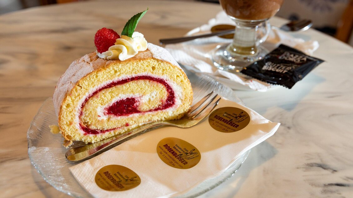Ein leckeres Stück Biskuitrolle mit einer Erdbeerfüllung, garniert mit Sahne und einer Erdbeere. Daneben steht ein Glas mit einem kalten Getränk und einige Servietten. | © TV Region Graz - Harry Schiffer