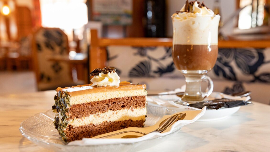Ein Stück Schokoladenkuchen mit Schichten und Sahne, serviert auf einem Teller. Dazu ein Glas mit einer kreativen Kaffeekreation und Sahne. | © TV Region Graz - Harry Schiffer
